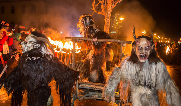 Krampuslauf v Mariazelle v noci: Trojica detailných a hrozivých Krampus masiek s kožuchmi v popredí, sprevádzajúca drevený vozík s plameňmi a ohňom, čo vytvára dramatickú atmosféru Behu čertov.