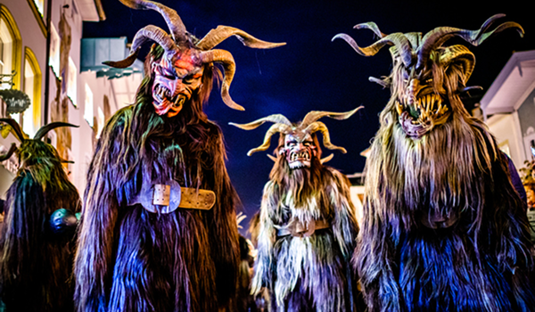 Štyri postavy Krampusov s rôznymi desivými maskami s rohmi a huňatými kožuchmi stoja tesne vedľa seba počas nočnej akcie.