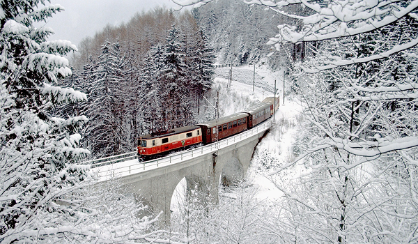 Mariazellerbahn (Mariazelská dráha): Historický červeno-biely vlak prechádza po viadukte cez hlboko zasneženú alpskú krajinu.