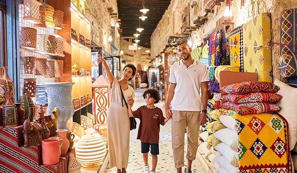 Rodina na prechádzke tradičným trhoviskom Souq Waqif v Dauhe, obklopená obchodmi s farebnými látkami, lampami a miestnymi remeselnými výrobkami.