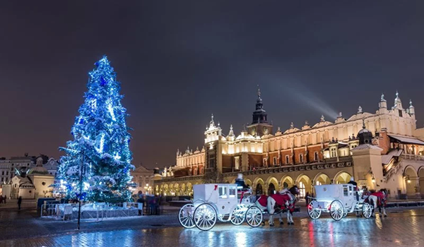 Nočný adventný zájazd do Krakova ukazuje Rynek Główny s obrovským modro osvetleným vianočným stromčekom a bielymi kočmi.
