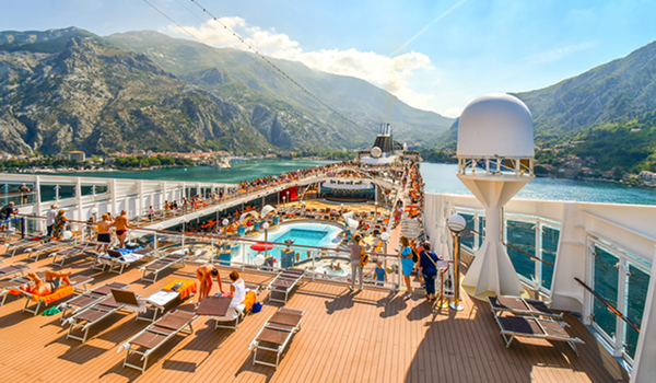Pohľad na palubu výletnej lode plnej turistov, ktorí odpočívajú pri bazéne a opaľujú sa s výhľadom na záliv a hornatú krajinu.