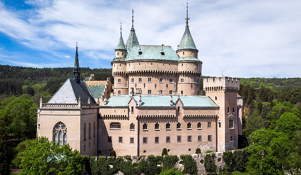Celkový pohľad na rozprávkový Bojnický zámok s ikonickými vežičkami a hradbami, týčiaci sa nad parkom počas slnečného dňa.