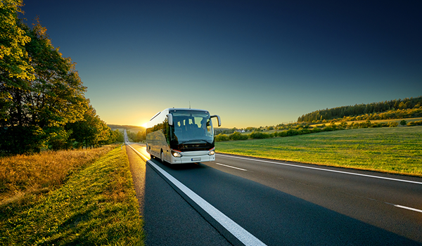 Luxusný autobus na dlhé cesty prechádza po vidieckej ceste lemovanej zeleňou pri jasnom západe slnka.