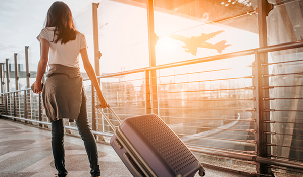 Žena s kufrom na kolieskach stojaca na letiskovom termináli, pozorujúca vzlietajúce lietadlo pri západe slnka.
