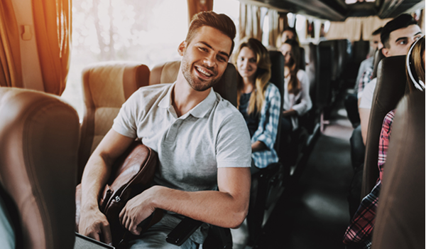 Usmievajúci sa mladý muž sediaci na sedadle vo vnútri autobusu plného cestujúcich, čo symbolizuje pohodlné cestovanie