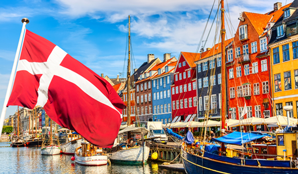 Pohľad na prístav Nyhavn s radom farebných domov a kotviacimi loďami, ktorému dominuje veľká dánska vlajka.