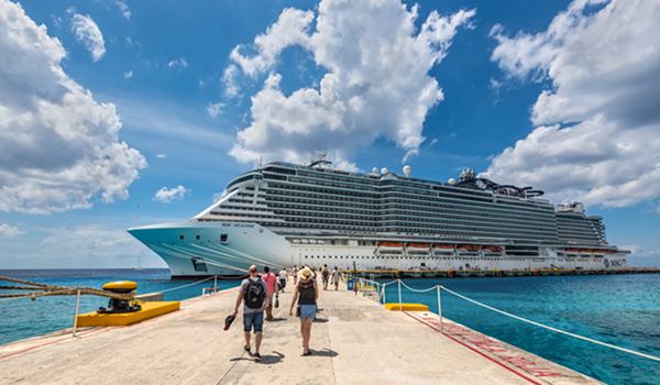 Pár kráča po betónovom móle smerom k obrovskej modernej luxusnej výletnej lodi kotviacej v prístave pod modrou oblohou.