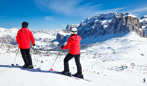 Dvaja lyžiari v červených bundách stoja na zasneženom svahu s nádherným výhľadom na hory.