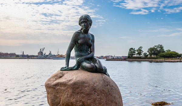 Slávna bronzová socha Malej morskej víly sediaca na kameni pri pobreží Kodane s prístavom a loďami v pozadí.