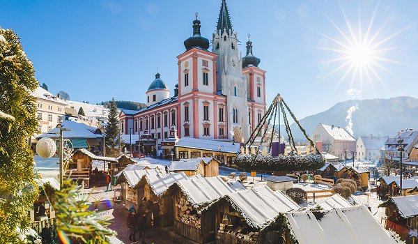 Adventné trhy v Mariazelle: Panoráma zasneženého námestia s drevenými stánkami, veľkým vencom a impozantnou Bazilikou Panny Márie.