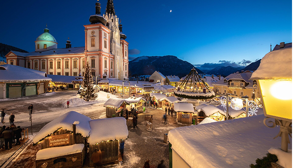 Nočné vianočné trhy v Mariazelle v zime: Osvetlené a zasnežené stánky pred historickou Bazilikou pri súmraku.