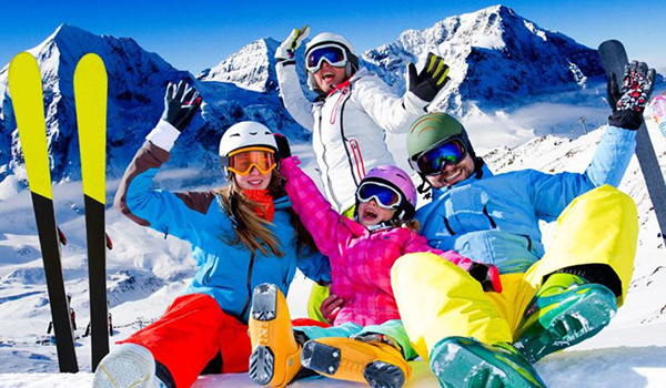 Rozosmiata rodina (rodičia a dve deti) sedí na snehu v lyžiarskej výstroji a radostne dvíha ruky, s lyžami zapichnutými v popredí.