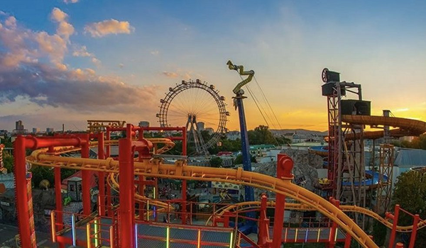 Zábavný park Prater vo Viedni pri západe slnka, zachytávajúci oranžovú horskú dráhu v popredí a ikonické Ruské koleso (Riesenrad) v pozadí.