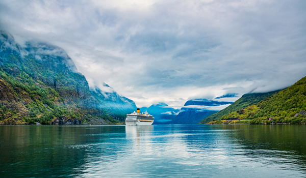 Obrovská výletná loď pláva po pokojnej vode medzi strmými, zelenými úbočiami v nórskom fjorde pod zamračenou oblohou.
