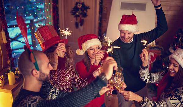 Skupina priateľov oslavuje Nový rok s vianočnými čiapkami, šampanským a prskavkami v útulnej horskej chate čo je ideálny Silvester na horách.