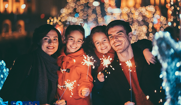 Rodina oslavuje Silvester počas zájazdu, drží prskavky a usmieva sa s jasným vianočným osvetlením v pozadí.