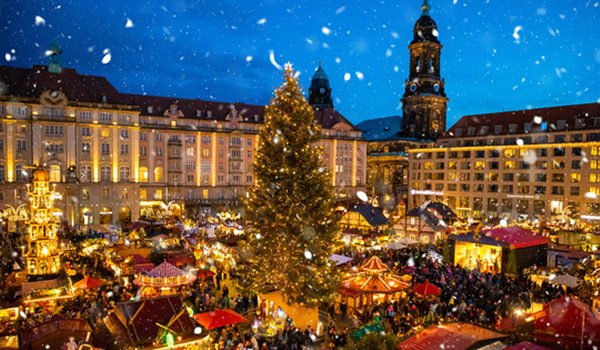 Nočný pohľad na tradičné vianočné trhy s vysokým, bohato osvetleným vianočným stromčekom a historickými budovami v pozadí, na ktoré padá sneh.