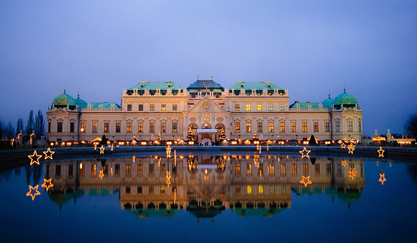 Osvetlený Horný Belvedere vo Viedni počas súmraku, odrážajúci sa v vodnej hladine pred palácom s vianočnou svetelnou výzdobou.