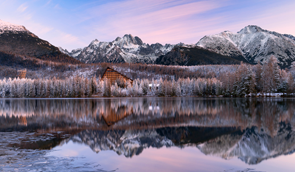 Panoráma zasnežených Vysokých Tatier s vrcholmi odrážajúcimi sa v pokojnej hladine jazera Štrbské pleso pri ružovom východe slnka.
