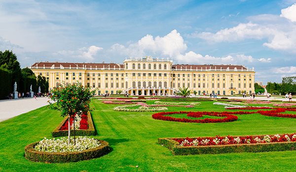 Slnečný pohľad na žltú fasádu zámku Schönbrunn vo Viedni s rozľahlými záhradami a farebnými kvetinovými záhonmi v popredí.